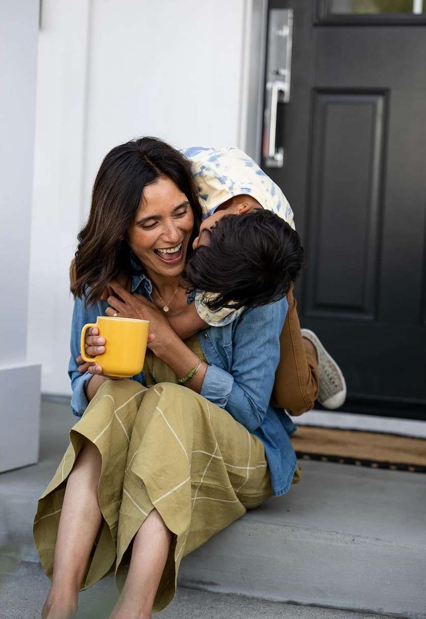 A happy mother and child embrace on a doorstep. The woman laughs while holding a yellow mug. A denim shirt adds a casual touch.
