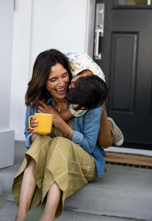 A happy mother and child embrace on a doorstep. The woman laughs while holding a yellow mug. A denim shirt adds a casual touch.