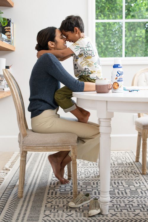 Woman and child with foreheads touching at a white table with Silk Almond Creamer, in a bright indoor kitchen setting with a patterned rug.