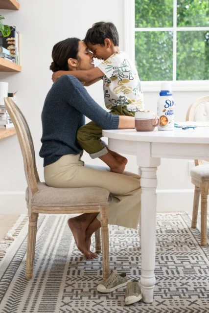 Woman and child with foreheads touching at a white table with Silk Almond Creamer, in a bright indoor kitchen setting with a patterned rug.