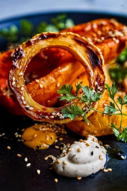 Close-up shot of roasted squash pieces, garnished with herbs and sesame seeds, on a dark plate. Food photography.