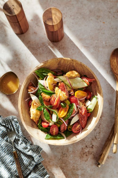 Overhead shot of fresh panzanella salad in a wooden bowl with serving utensils and wooden salt and pepper shakers, sunlit.