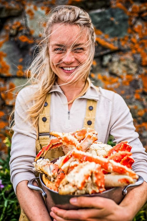 Blonde woman smiles holding a pot full of cooked king crab legs in front of a lichen-covered stone wall in an outdoor setting.