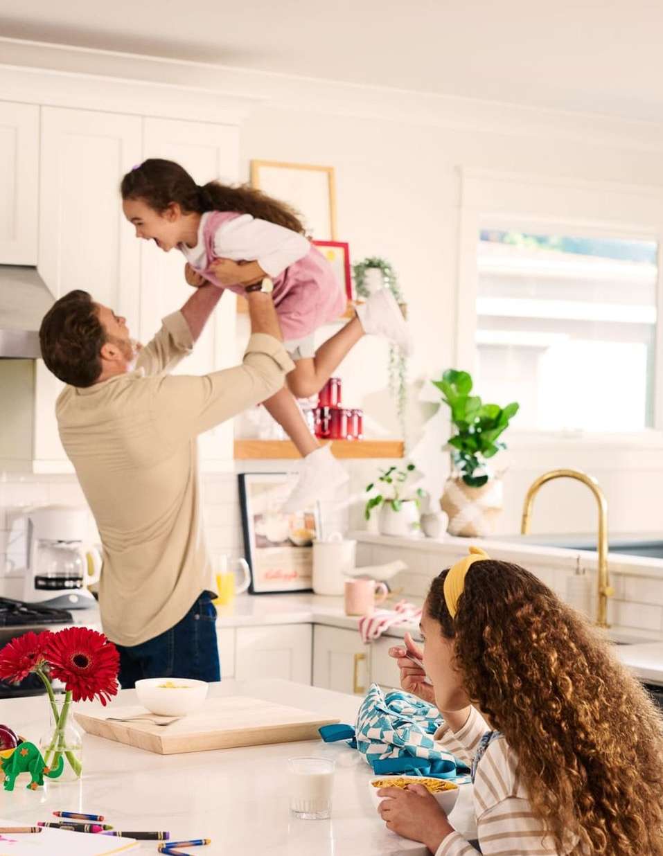 Father lifts his daughter in the air while another daughter eats at the counter in a well-lit, white kitchen with red and green accents.