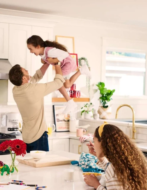 Father lifts his daughter in the air while another daughter eats at the counter in a well-lit, white kitchen with red and green accents.