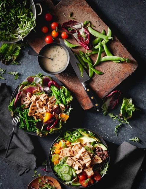 Overhead shot of two bowls of salads with grilled chicken, fresh vegetables, and herbs, plus tomatoes, snap peas, and dressing.
