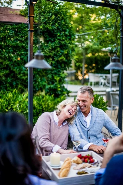 A smiling couple sits at an outdoor table with a tray of snacks, drinks, and a candle, surrounded by greenery.