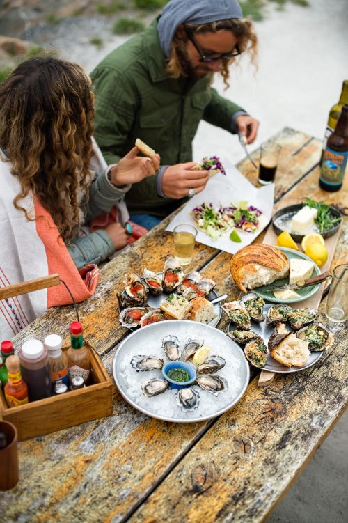 Two people at a wooden table outdoors, enjoying oysters, bread, and drinks, with various plates and bottles arranged on the table.
