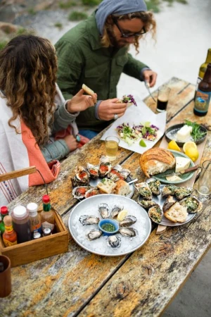 Two people at a wooden table outdoors, enjoying oysters, bread, and drinks, with various plates and bottles arranged on the table.