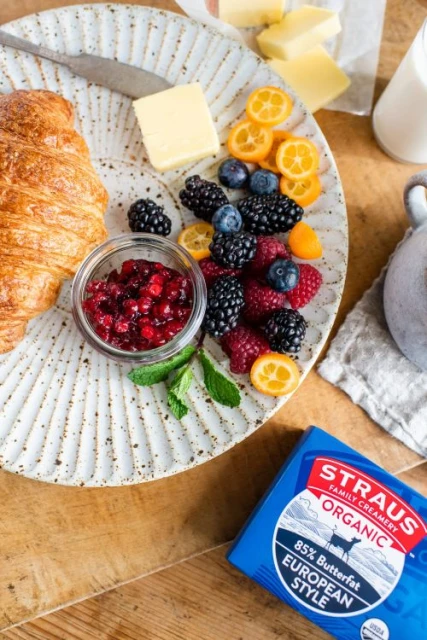 Overhead shot of a plate with a croissant, butter, fruit, and jam, alongside a butter package on a wooden table, bright, natural light.