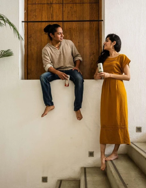 Barefoot man and woman in casual clothes hold containers indoors on stairs near a wood door and houseplants in natural light.