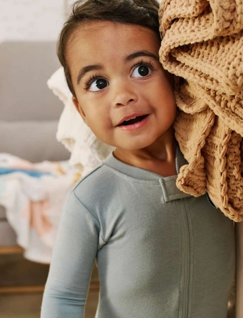 Adorable toddler in grey pajamas peeks out playfully from behind a textured, beige blanket, looking up with wide, expressive eyes.