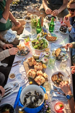 Overhead shot of a group of people dining outdoors at a table filled with mussels, bread, salad, drinks, and various dishes.