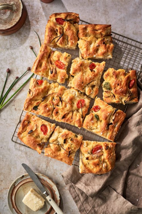 Overhead shot of focaccia bread cut into squares, seasoned with tomatoes and herbs on a wire rack next to a butter knife and butter.