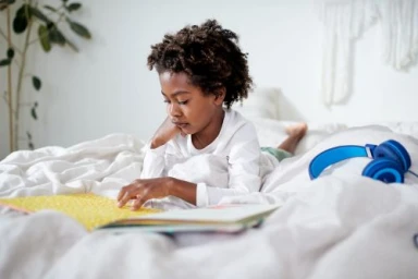 Young Black child reading a book while lying on a white bed with blue headphones nearby, captured by Dawn Heumann