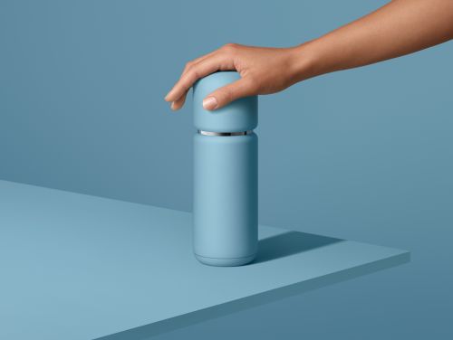 Close-up studio shot of a hand on a light blue reusable bottle against a light blue background, emphasizing product design and color.