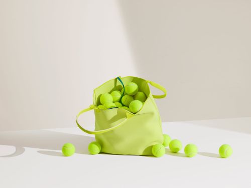 Lime green tote bag filled with tennis balls scattered on white surface in studio lighting. Minimalist sport equipment.
