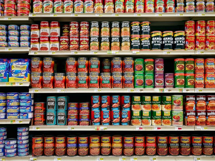 Canned goods like beans, chili, and tuna stacked on shelves at a grocery store. Rows of cans are organized by brand and variety.
