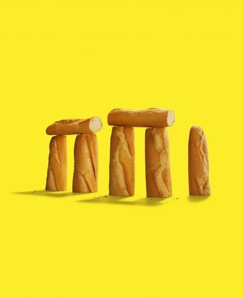 Stonehenge made of bread baguettes on a bright yellow background, creating a whimsical and surreal food composition.
