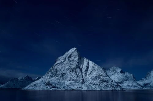 Snow-covered mountains reflecting in the water under a dark blue night sky with visible stars and star trails in a scenic landscape.