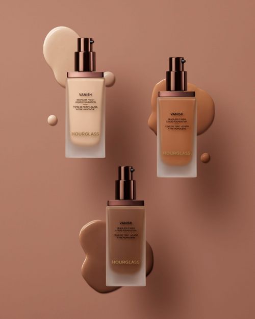 Three bottles of Hourglass Vanish foundation in different shades arranged with matching foundation smears on a neutral background.