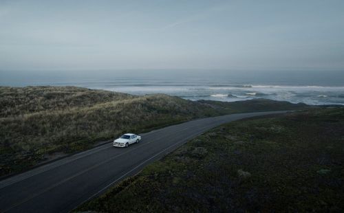 A white sedan drives along a coastal highway under a cloudy sky, with ocean waves visible in the background.