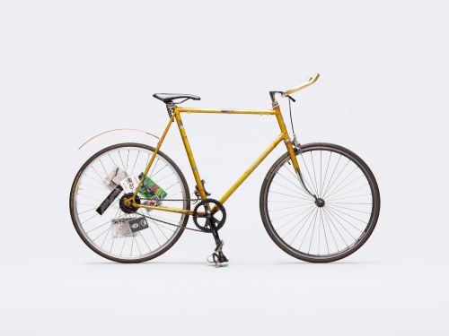 A yellow bicycle stands against a solid white background, featuring a classic frame and attached papers on the rear wheel.
