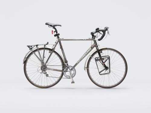 Silver Motobecane bicycle with accessories, including a rear rack, fenders, and front carrier, standing against a neutral gray background.