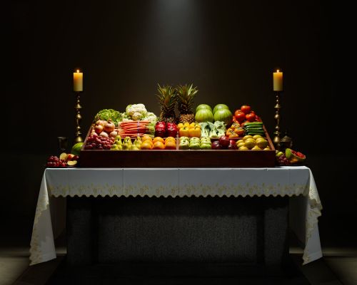 Still life photograph of a table laden with a colorful assortment of fruits and vegetables, flanked by lit candles, against a dark background.