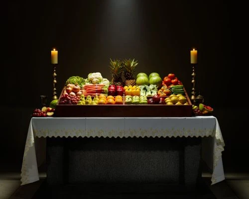 Still life photograph of a table laden with a colorful assortment of fruits and vegetables, flanked by lit candles, against a dark background.
