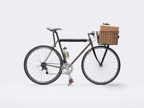 A bicycle with a brown frame is carrying a cardboard box in a wire basket. A water bottle is attached to the bike.
