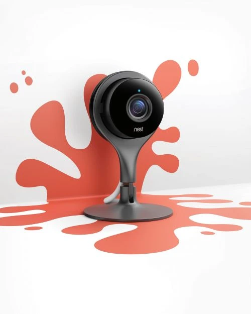 Nest security camera stands in front of white wall featuring coral paint splatters; product shot in a brightly lit interior.