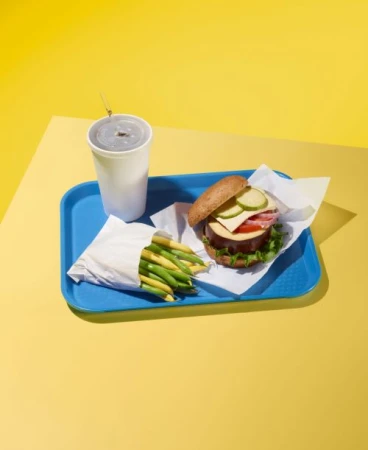 Burger with green and yellow beans, and a drink, served on a blue tray against a yellow background. Fast food still life.
