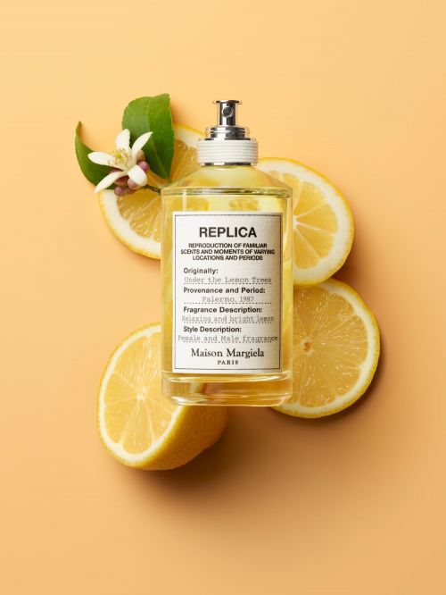 Close-up studio shot of Maison Margiela REPLICA fragrance surrounded by fresh lemon slices and green leaves on a peach background.
