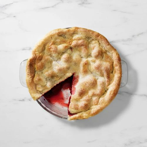 Overhead shot of a baked pie with a slice cut out, revealing a red fruit filling in a clear glass baking dish on a marble surface.