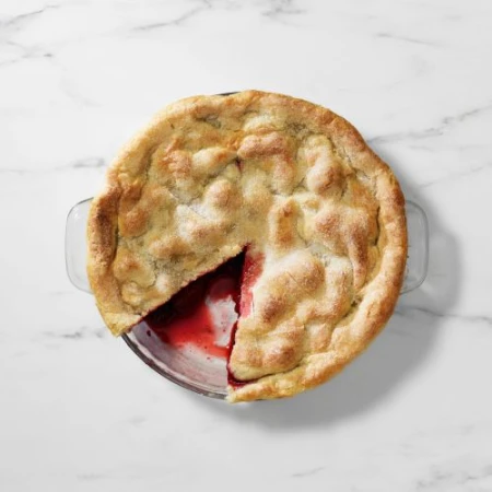 Overhead shot of a baked pie with a slice cut out, revealing a red fruit filling in a clear glass baking dish on a marble surface.