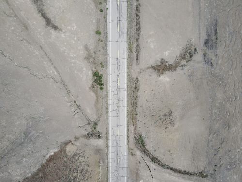 Aerial view of a cracked road running through a desolate, arid landscape with sparse vegetation and dry earth. The road appears worn.
