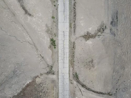 Aerial view of a cracked road running through a desolate, arid landscape with sparse vegetation and dry earth. The road appears worn.