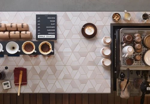 Overhead shot of a coffee preparation station with coffee bags, cups, a grinder, and an espresso machine on a patterned tile counter.