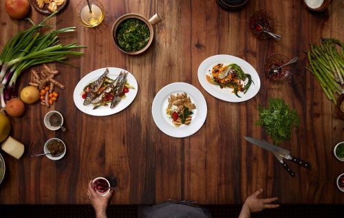 Overhead shot of various dishes and ingredients arranged on a dark wood table, capturing a culinary preparation scene.