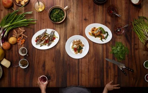 Overhead shot of various dishes and ingredients arranged on a dark wood table, capturing a culinary preparation scene.