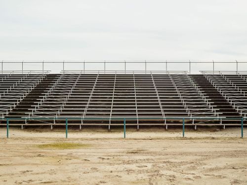 Wide shot of rows of empty metal bleacher seats at an outdoor venue beneath a cloudy sky, overlooking a dirt ground with tracks.
