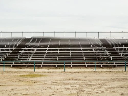 Wide shot of rows of empty metal bleacher seats at an outdoor venue beneath a cloudy sky, overlooking a dirt ground with tracks.