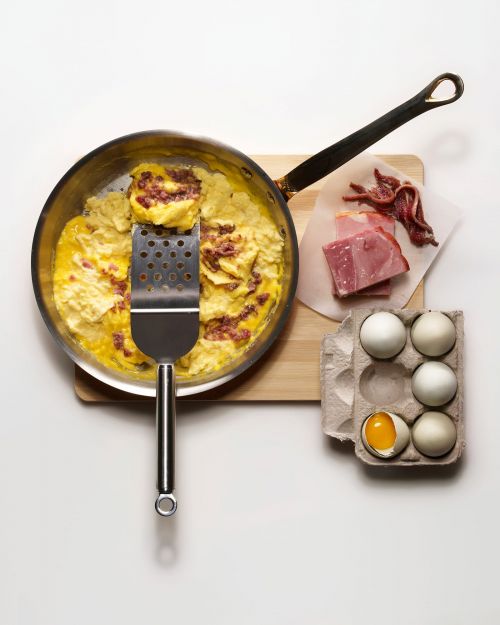 Overhead studio shot of a skillet with an omelet and spatula, with ham and eggs beside, on a white surface and a wooden cutting board.