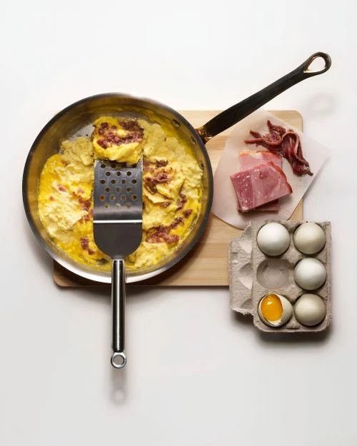 Overhead studio shot of a skillet with an omelet and spatula, with ham and eggs beside, on a white surface and a wooden cutting board.
