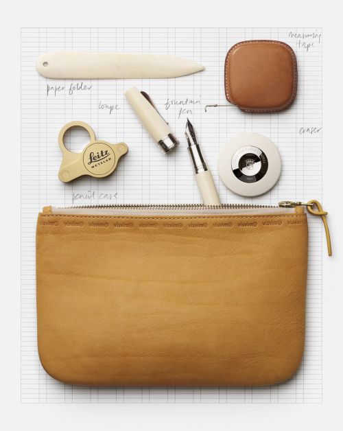 Still life of leather pouch, fountain pen, paper folder, and measuring tape on a light gray gridded surface, overhead shot.
