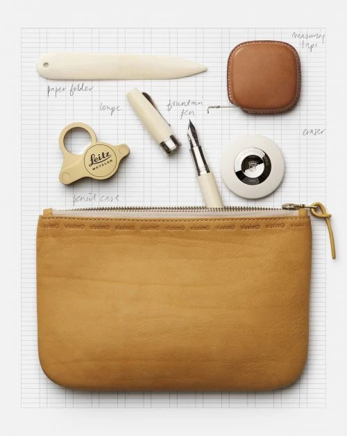 Still life of leather pouch, fountain pen, paper folder, and measuring tape on a light gray gridded surface, overhead shot.