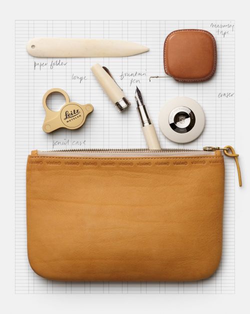 Close-up still life featuring a tan-colored leather pouch, writing instruments, and office supplies arranged on a grid background.