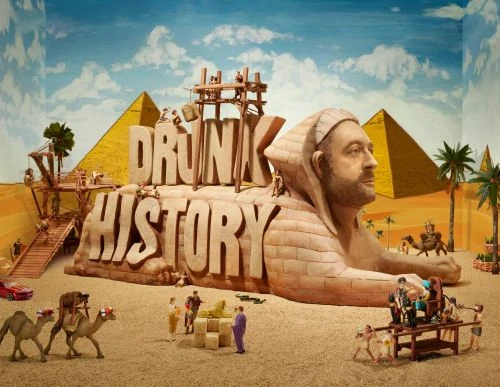 Whimsical diorama of an Egyptian scene with a sphinx-like sculpture, pyramids, palm trees, and miniature figures, creating a humorous historical tableau.