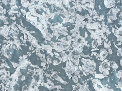 Aerial view of a glacier featuring abstract patterns of white ice and grey water in a natural environment, taken with an overhead angle.